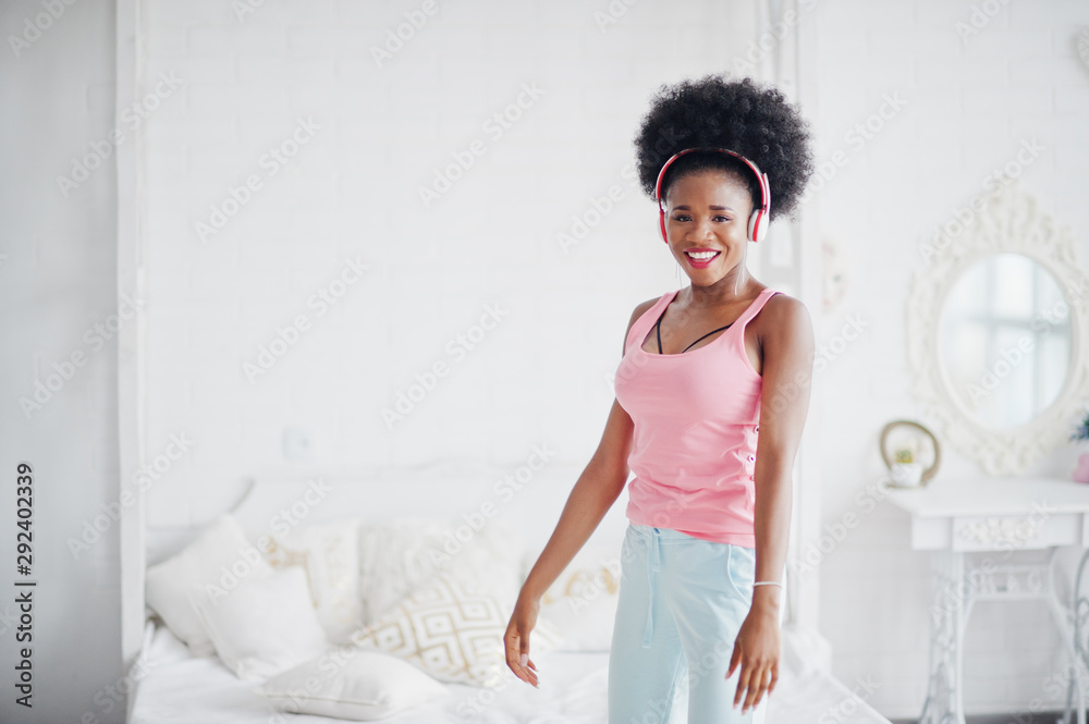 Young african american woman in pink singlet dancing and listen music on earphones at her white room. Young african american woman in pink singlet dancing and listen music on earphones at her white room.