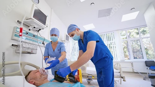 Male unconscious patient laying on bed and two doctors trying to save life with modern technologies. Male specialist using defibrillator to resuscitate patient in new emergency room.