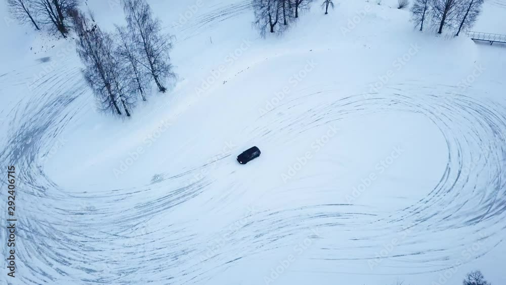 A black car practices race drifting in the snow  on frozen lake around trees of the road course. A steady overhead following shot of a car drift racing a road course