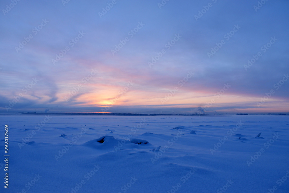 View on winter snowy river with snowbanks and blurred background with sunset, shore front and town chimney. Copy space