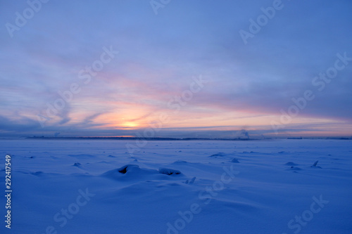 View on winter snowy river with snowbanks and blurred background with sunset  shore front and town chimney. Copy space