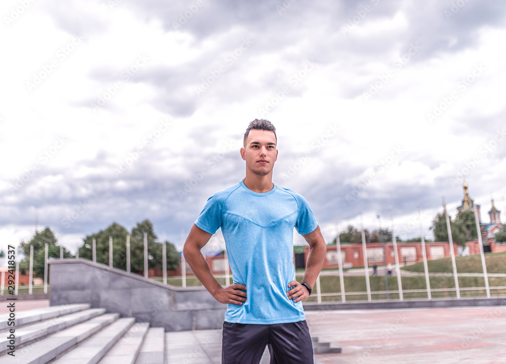 Confident and strong male athlete, summer city, trainer stares intently, background is blue clouds, free space for motivation text, fitness workout workout. Sportswear t-shirt, fitness bracelet.