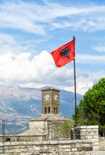 The clock tower and Abanian flag on Gjirokaster Fortress Castle with mountains and blue sky with white clouds in background.