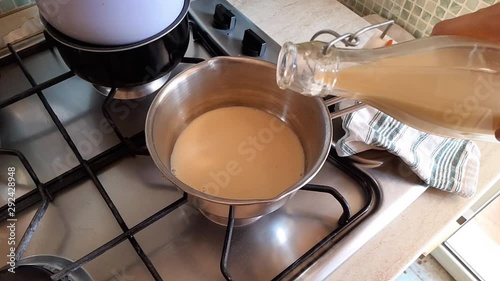 Pouring home made plant milk from glass bottle in a steel pan put on the cooker for breakfast latte. Slow motion
