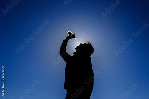 Backlit sportsman drinking water after exercising. Blue sky background.