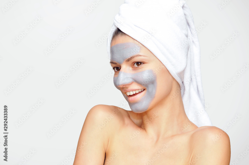 Beautiful young woman with a gray cosmetic mask on her face and a white towel on her head with bare shoulders. Beautiful young woman with a gray cosmetic mask on her face and a white towel on her head with bare shoulders.
