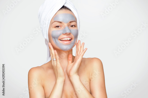 Beautiful young woman with a gray cosmetic mask on her face and a white towel on her head with bare shoulders.
