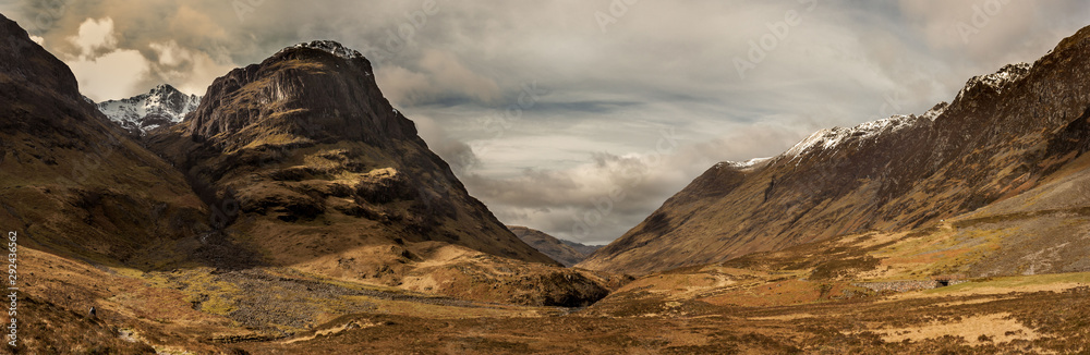 A panoramic view of the valley of Glencoe, in the Highlands of Scotland, with the mountain peaks covered in snow and a dramatic cloudy sky