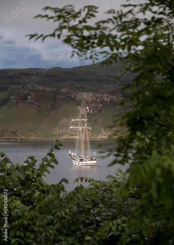 Vertical landscape of a white sailing boat off the coast of Potree, in Scotland, framed by blurred green foliage in the foreground and with tall cliffs in the background. Isle of Skye. UK. Seascape photo