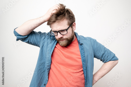 Quadro su tela Young bearded man with glasses is in doubt, he is confused, looks down thoughtfully and scratches his head, on a white background