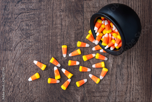 Black ceramic cauldron filled with holiday candy corn tipped over on a rustic wood background, spilling over onto the background photo