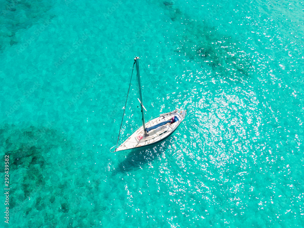 Fototapeta premium High angle aerial drone bird's eye view of a sailing yacht with sails lowered anchored in shallow, turquoise water in the Atlantic Ocean near the coast of Fuerteventura, Canary Islands, Spain, Europe.