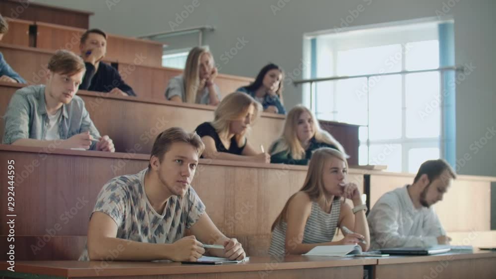 Modern students, men and women, sit at desks in a large classroom and write a lecture notes.