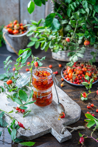 Rose hip jam in jar pots on wooden board. Ripe red berries with leaves in plate and basket. Autumn harvesting season, fresh vitamins on gray background. Healthy nutrition, homemade marmalade concept
