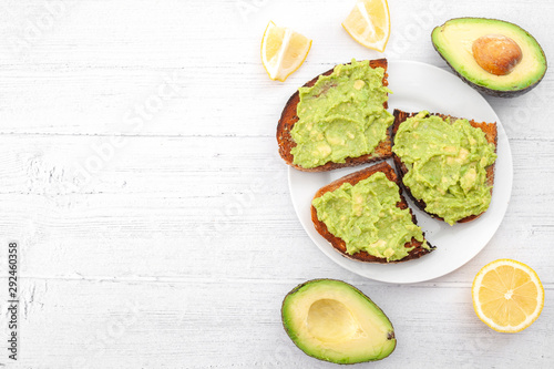 Healthy fat diet and mexican food conceptual idea with smashed avocado into guacamole spread on rye bread toast surrounded by lemon and avocados isolated on white wood table with copyspace