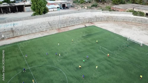 Drone footage of two teams playing at a football field. Aerial view of a soccer court and players running after the ball on a match with buildings on the background. Birds eye of pitch from above.