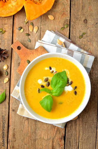 Bowl of cream pumpkin, carrot soup with fresh basil, pumpkin seed on rustic wooden table. Top view, vertical photo.