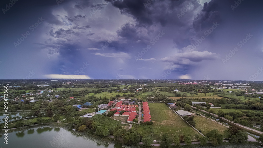 Aerial view above the lake, village and green rice fields plantations, view of heavy raining and storm with dark clouds moving in the sky background, Krajub Reservoir, Ban Pong, Ratchaburi, Thailand.