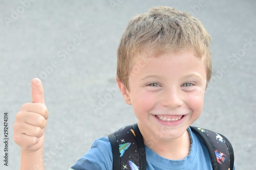 Blonde kid with blue eyes wearing a backpack giving thumbs up