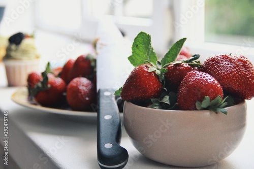 Fresh tasty and delicious strawberries in a white bowl on a window background. Healthy nutrition and food concept, vitamins for autumn's health. Bright colors. Berries with knife against cupcakes.