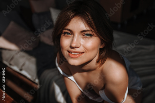 The girl sits on the bed in the sunlight and looks at the camera and smiles close-up