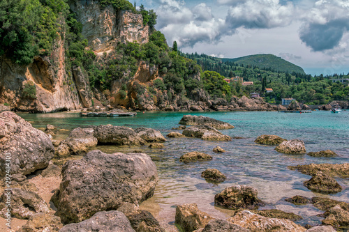 Seascape – lagoon with turquoise water, mountain with cliffs, green trees, bushes, rocks in a blue water and clouds on the sky. Corfu Island, Greece. 