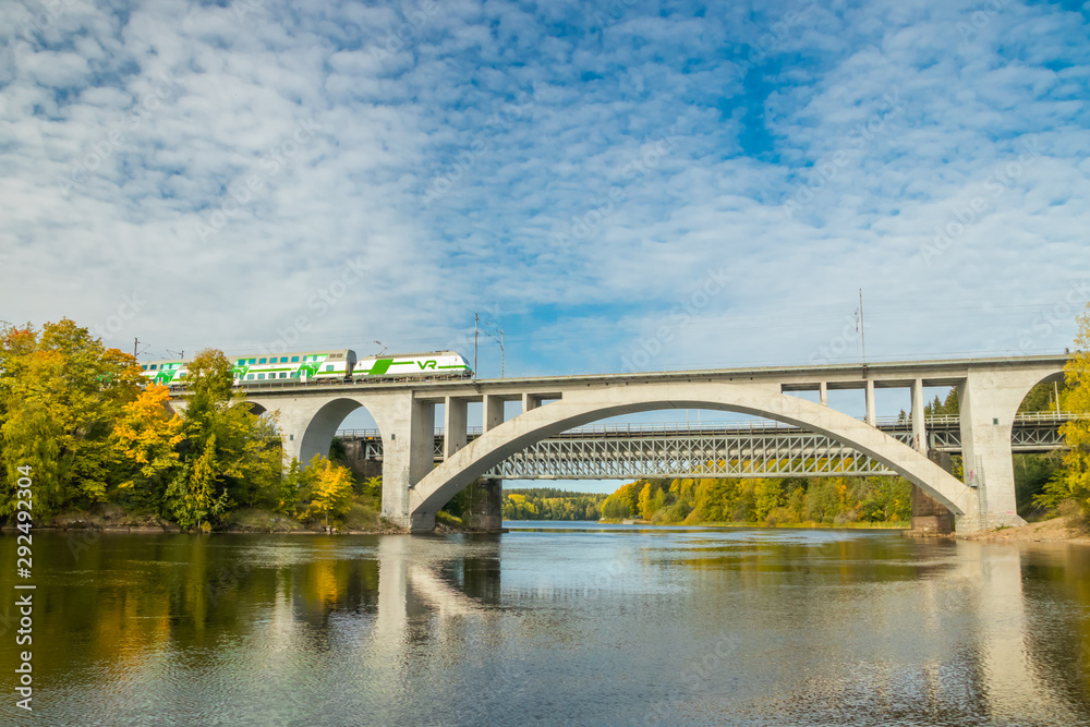Fototapeta premium Kouvola, Finland - 25 September, 2019: Autumn landscape of bridge with moving passenger train and Kymijoki river waters in Finland, Kymenlaakso, Kouvola, Koria