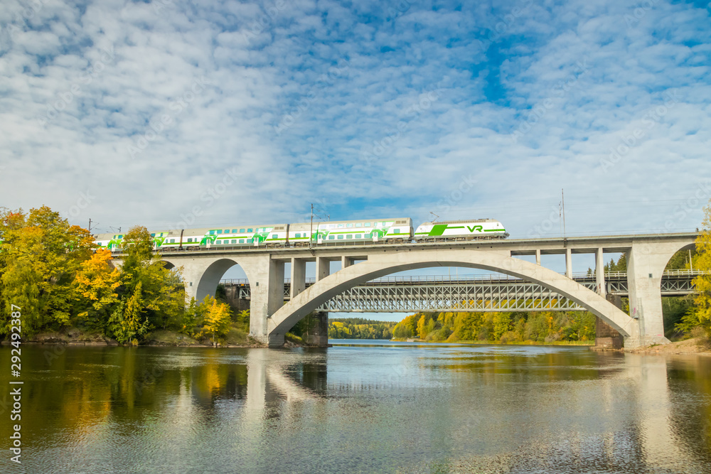 Naklejka premium Kouvola, Finland - 25 September, 2019: Autumn landscape of bridge with moving passenger train and Kymijoki river waters in Finland, Kymenlaakso, Kouvola, Koria