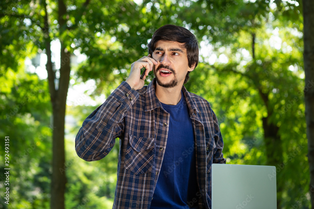 Young brunet freelancer with beard and mustache looks surprised sitting with a laptop in a park and speaking on the smartphone. Work at any place Young brunet freelancer with beard and mustache looks surprised sitting with a laptop in a park and speaking on the smartphone. Work at any place