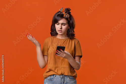 Pretty brunette woman in a t-shirt and beautiful headband is displeased checking smartphone for notifications isolated over orange background. Daily communication