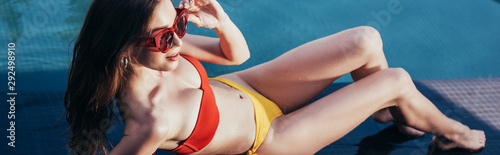 panoramic shot of attractive young woman in sunglasses relaxing near swimming pool