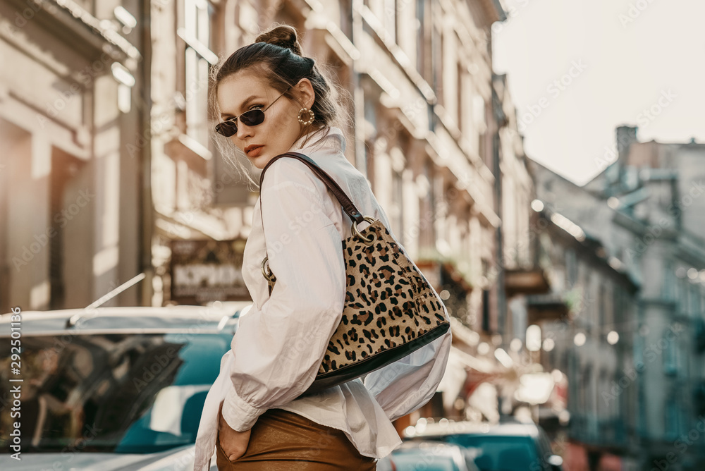 Outdoor autumn fashion portrait of elegant, luxury woman wearing sunglasses, trendy white shirt, leather trousers, with animal, leopard print bag, walking in street of European city. Copy, empty space - obrazy, fototapety, plakaty 