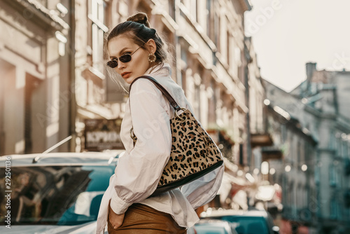 Outdoor autumn fashion portrait of elegant, luxury woman wearing sunglasses, trendy white shirt, leather trousers, with animal, leopard print bag, walking in street of European city. Copy, empty space