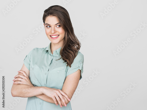 Cheerful young woman portrait. Smiling girl with crossed arms looking away. Isolated on grey background