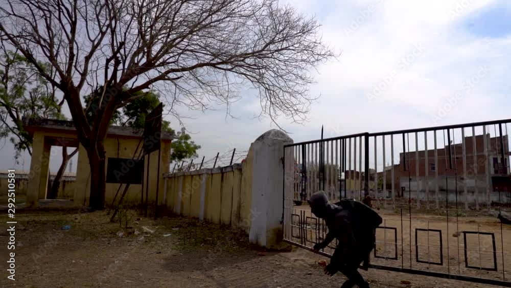 A burglar entering a deserted empty building area by jumping off the gate and running with covered face and bag on back