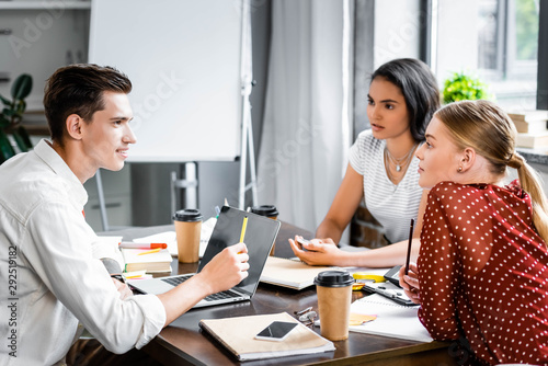 three multiethnic friends sitting at table and talking in apartment