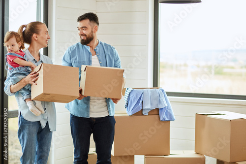 Bearded dark-haired man wear blue shirt enter house and keep box, fair-haired woman wear blue shirt keep boxes and child. They look at each other. Boxes and big window in background © alfa27