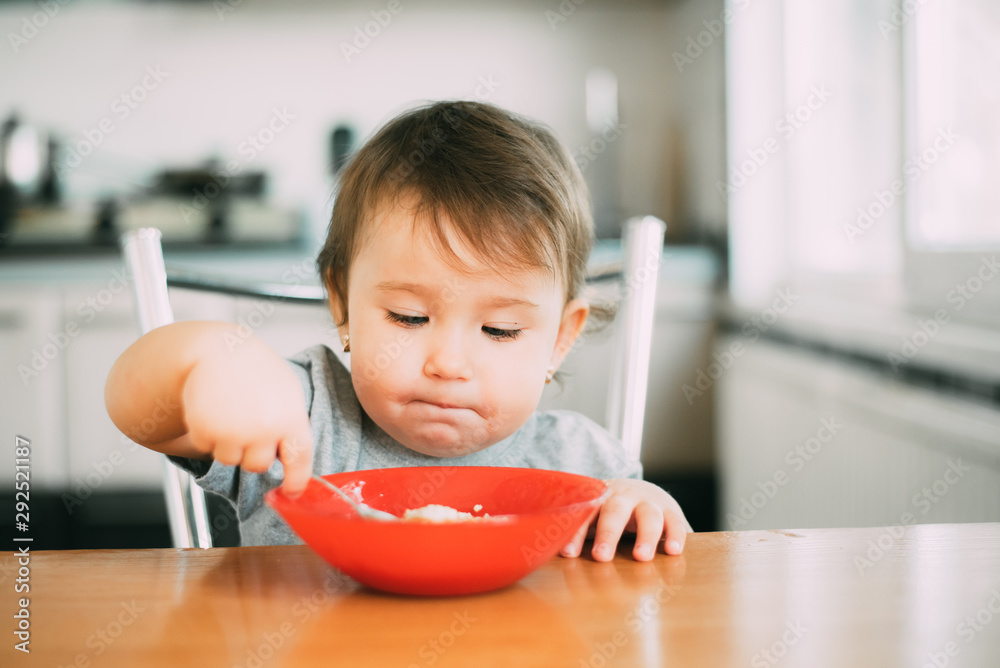 little girl child in the kitchen on their own eating oatmeal with a red plate little girl child in the kitchen on their own eating oatmeal with a red plate