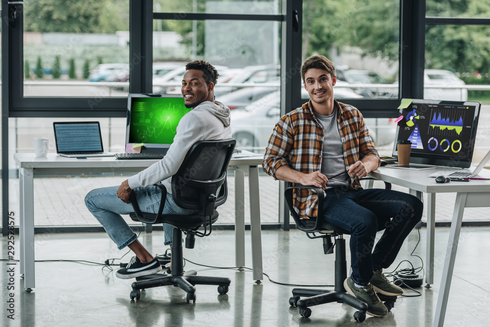 two young multicultural programmers looking at camera while sitting near computer monitors with graphs and charts on screen two young multicultural programmers looking at camera while sitting near computer monitors with graphs and charts on screen