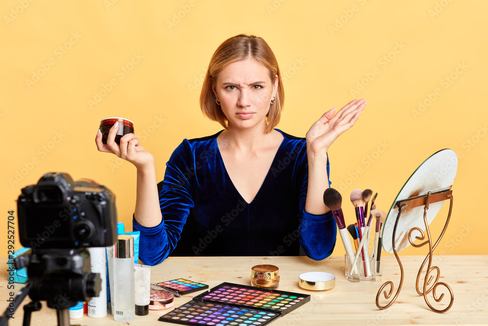 Isolated shot of displeased beauty blogger girl spreads hands, holds new moisturizing face cream, frowns face in dislike, dissatisfied with quality of the product. Video blogging concept. Isolated shot of displeased beauty blogger girl spreads hands, holds new moisturizing face cream, frowns face in dislike, dissatisfied with quality of the product. Video blogging concept.