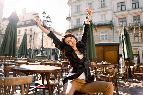 beautiful woman sitting in a cafe on a city street, fashion style, hands up, wake up in the morning, enjoy sunny day