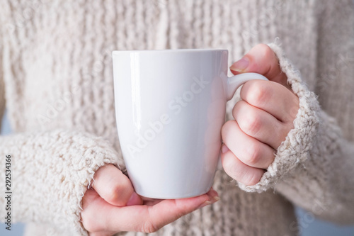 Woman in warm sweater is holding white mug in hands