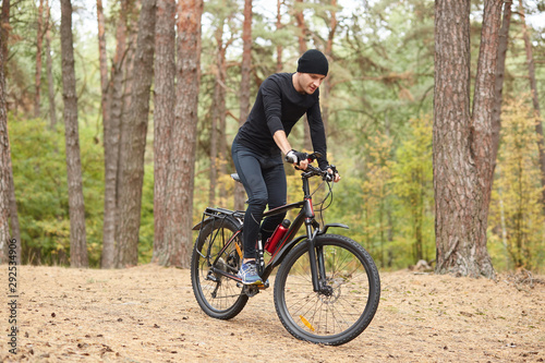 Photo of cyclist on forest trail, trip on bikes in lush green nature, young attractive guy wearing black sportwear rides around trees in forest, spends free time in open air. Healthy lifestyle concept