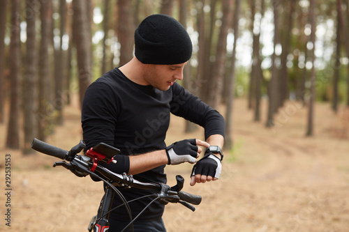 Outdoor shot of handsome man using his smart watch, riding bike in forest, wearing black sportwear and cap, looks athis device, spending leisure time inopen air, enjoys fresh air and beautiful nature. photo