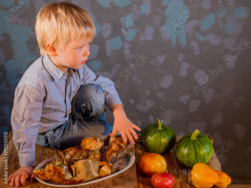 little blond child sits at table in front of dish of fried fish. kid comes up with entertainment and actively masters necessary skills. Yellow tomato  sweet pepper  pumpkins. Hands close up