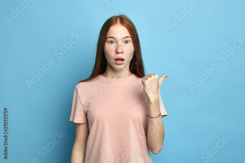 beautiful emotional freckled girl with ginger hair, has surprised expression, indicates aside, shows free space for advertisement.isolated blue background, studio shot. close up portrait