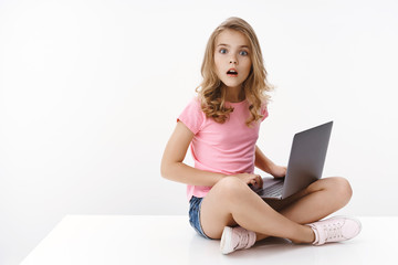 Amazed excited smart cute european little girl, sit crossed legs laptop, studying online e-learning, turn away pc screen stare camera thrilled, explain awesome cartoon episode, white background
