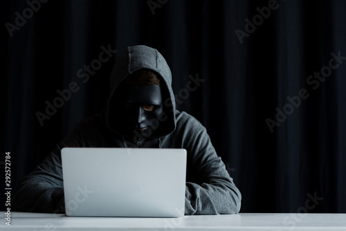 anonymous internet troll in mask typing on laptop keyboard on black