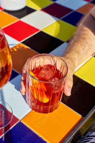 Male hand holding a glass of Negroni cocktail on a hot sunny day. Colorful tiled table photo