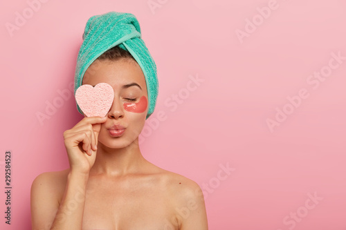 Isolated shot of pretty female model has eyes closed, lips rounded, stands with bare shoulders, enjoys beauty treatments, holds cosmetic sponge over eye, cares about face wears hydrogel patches, towel